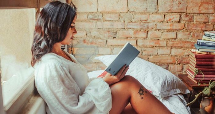 a tanned skin woman sitting upright in a bed and reading a book.jpg.optimal