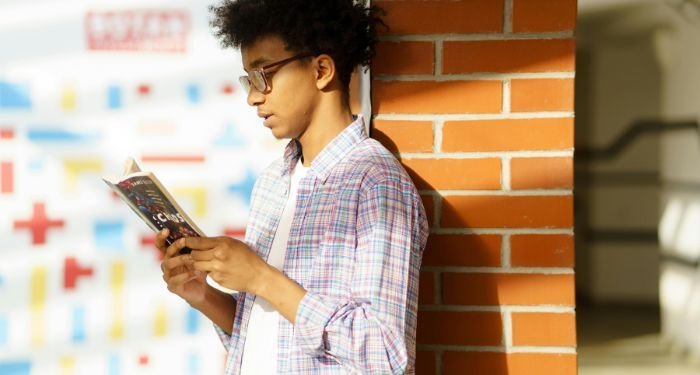 brown skinned Black man with afro reading a book against a brick wall.jpg.optimal