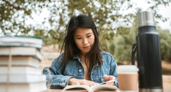 light skinned Asian woman reading at a table with a thermos and a coffee cup.jpg.optimal