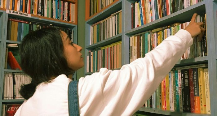 a light brown skinned woman with dark hair reaching for a book in a used bookstore.jpg.optimal