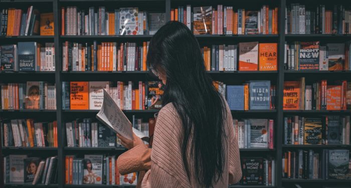 asian person reading a book in front of a filled bookshelf in a book store.jpg.optimal